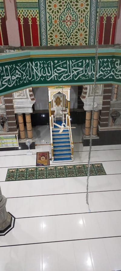 An interior shot of a mosque reveals a raised pulpit with blue carpeted steps. Green prayer rugs line the tiled floor leading to it. Ornate Arabic calligraphy borders the upper section of the mosque. Pillars with detailed ornamentation flank the pulpit's entrance. A ceiling fan hangs nearby. A hanging chain and the top of a pillar are visible at the bottom edges of the image.