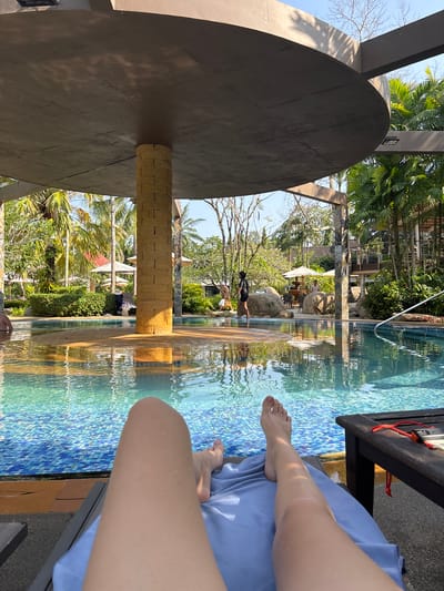 The image shows a scene at a swimming pool in Khok Kloi, Thailand. A person is sitting on a lounge chair next to the pool, visible from the knees down. The person is draped with a blue cloth. The pool has a blue tile mosaic and reflects the surrounding foliage, including palm trees and other greenery. A structure with a large, circular overhanging roof provides shade. A person in a black suit is walking by the pool and there is a patio area in the background.