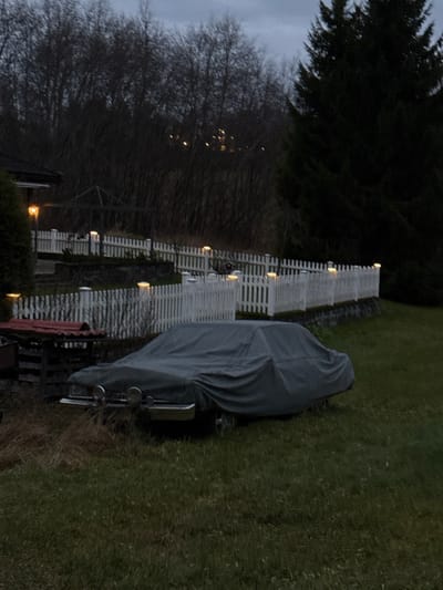 The image depicts an outdoor residential scene in Jessheim, Norway, observed during low light conditions, suggesting dusk or early night. In the foreground, a vehicle is parked on a grassy lawn, almost entirely covered by a grey tarpaulin. The front of the vehicle is partially visible beneath the cover, revealing a chrome bumper, a grille, and two circular auxiliary lights. To the left of the covered car, a small, dark wooden structure with a reddish, possibly tiled, roof is present.

In the midground, a white picket fence with multiple illuminated post-cap lights extends horizontally, then ascends a gentle slope, forming a tiered barrier for what appears to be a terraced garden area. An outdoor lamp with warm illumination is visible on the corner of a building, likely a house, located in the upper-left quadrant. The background features a dense cluster of bare deciduous trees, with indistinct, scattered lights visible through their branches, suggesting a distant populated area. A large, dark coniferous tree occupies the upper right portion of the background. No individuals or active subjects are present in the scene.