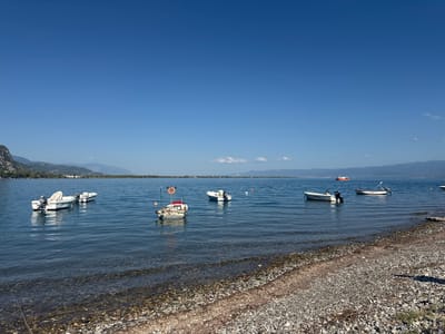 The image shows a body of water, likely a bay or lake, with several small boats anchored or moored near the shore. The water is calm and reflects the clear blue sky. The shoreline is composed of pebbles and a small amount of vegetation. In the background, there are rolling hills and mountains under a cloudless sky. The scene appears to be a tranquil coastal or lakeside setting. Based on the scenery, the location is likely the Municipal Unit of Kamena Vourla, Greece, known for its coastal geography and mountainous backdrop.