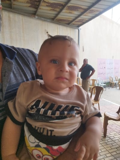 The image captures a scene in Idhna, Palestinian Territory, under a corrugated metal canopy. In the foreground, a light-skinned infant with blue eyes and light hair is prominently featured, held by an adult whose torso and left shoulder, clad in a striped dark shirt, are visible on the left. The infant wears a light brown long-sleeved shirt with "NIKE" in black lettering across the chest, and a colorful cartoon-patterned undershirt or bib is visible below. The child's gaze is directed forward. In the midground, to the right of the infant, a bearded adult male, wearing a dark green polo shirt and light-colored trousers, stands with his right hand raised near his chin and his left hand on his hip. He appears to be looking towards the left. Behind this man and extending across the midground are multiple rows of light brown and white plastic chairs arranged on a tiled surface. A white sheet or banner adorned with Arabic script is affixed to the light-colored wall in the background. The overhead corrugated metal canopy shows signs of rust. The ground is paved with rectangular tiles in shades of reddish-brown and tan.