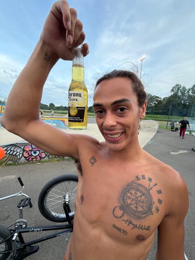 A shirtless, light-skinned man is holding a bottle of Corona Extra beer in his left hand at what appears to be a skate park. The beer label is legible. He is smiling and looking at the camera. On his left wrist, there is a tattoo that reads "557". On his left shoulder, there is a compass tattoo and the words "Hayse Wall" and "Faneto." There is a BMX bike visible behind him on the right. Graffiti art is visible on the ground. In the background, there are people on the skate park surface, trees and a light post. The location is Kettering, United Kingdom.