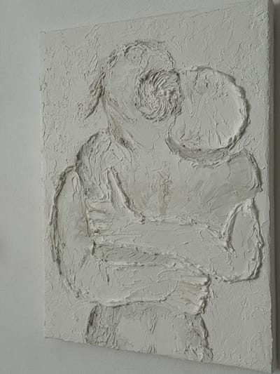 This is a close-up, angled shot of a textured white bas-relief sculpture. The sculpture depicts two figures embracing, with one figure's head tilted towards the other. The relief is rendered with thick, impasto strokes, creating a highly tactile surface. The background is a plain, textured white wall, suggesting the sculpture is displayed indoors. The image does not contain any information about its location in Ivatsevichy, Belarus.