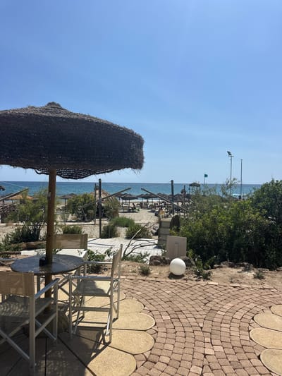 This image captures a serene beachside scene in Biddeputzi/Villaputzu, Italy, under a clear blue sky. In the foreground, a thatched umbrella shades a round wooden table accompanied by two white director-style chairs. The table has a small ashtray on it. The seating area is situated on a paved patio made of tan, irregularly shaped stones, bordered by a cobblestone path.
Beyond the immediate seating area, the sandy beach stretches out towards the vibrant blue sea. Numerous beach umbrellas and loungers are visible, suggesting a public beach with amenities. Lush green bushes and trees are scattered along the shoreline and further inland, adding a touch of nature to the scene. A few people can be seen relaxing on the beach in the distance.
The overall impression is one of a tranquil summer day at the beach, ideal for relaxation and enjoying the coastal Italian atmosphere.