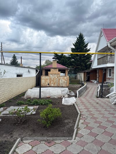 The image depicts an outdoor scene in Druzhino, Russia, characterized by a cloudy sky. A two-story house with a red-tiled roof is visible in the background, alongside a tall conifer tree. A brick barbecue structure with a red roof is positioned in the midground. To the right, a paved walkway with a red and gray pattern leads towards the house. A garden bed filled with plants and some white bags is located beside the walkway. A yellow gas pipe runs vertically near the house. In the foreground, a low white building with a textured exterior is present.