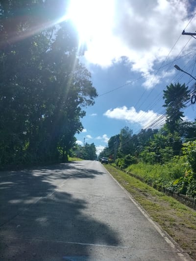 The image depicts a road in Kidapawan City, Philippines, on a sunny day with scattered clouds. Lush green trees line the left side of the road, with their leaves casting dappled shadows on the asphalt. On the right side, the road is bordered by a grassy embankment and more trees, with utility poles and a network of electrical wires overhead. A dark-colored SUV is parked on the side of the road further down. The sky is a vibrant blue, with a bright sunburst effect emanating from the upper left corner, suggesting it is midday or early afternoon. The road appears to be a two-lane asphalt surface with white lane markings.