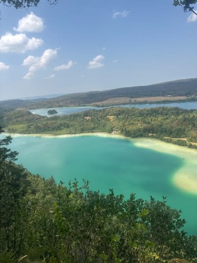 The image captures a panoramic view of a vibrant turquoise lake set against a backdrop of rolling, tree-covered hills and a clear blue sky with scattered white clouds. In the foreground, lush green foliage partially obscures the view, adding depth to the scene. The lake's surface exhibits a gradient of colors, transitioning from a deep turquoise in the foreground to a lighter, almost milky hue near the shoreline, suggesting shallows or submerged sediment. Further in the distance, another body of water is visible, with a small, tree-covered island nestled within it. The landscape appears serene and natural, indicative of a rural or park-like setting. The location is identified as the Lac de Chalain, situated in the commune of La Chaux-du-Dombief, France.