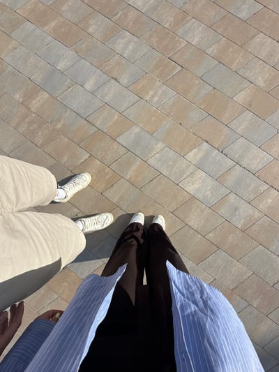 Two people are standing on a brick-paved ground in what appears to be a public space, possibly in Peguera, Spain. The person on the left is wearing beige shorts and white sneakers with white socks. The person on the right is wearing brown pants, white sneakers, and a light blue striped shirt. The background is a repeating pattern of rectangular paving stones. The lighting suggests a sunny day.