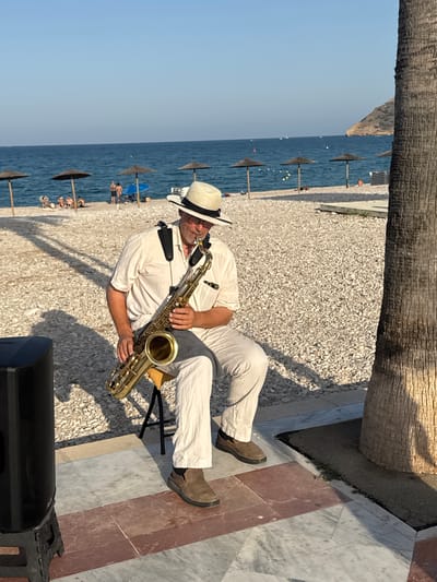 A man is sitting on a small stool on a pebbled beach in l'Alfàs del Pi, Spain, playing a saxophone. He is wearing a white straw hat, glasses, a light-colored short-sleeved shirt, and light-colored pants. A black speaker is positioned to his left, and a palm tree trunk is visible on the right side of the frame. In the background, the blue sea stretches to the horizon, with a beach dotted with umbrellas and a few people visible in the distance. The sky is clear and blue. The ground in the foreground is paved with marble and red stone tiles.