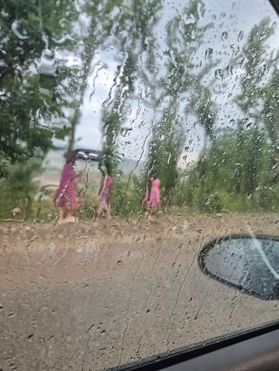 Through a rain-streaked car window, two figures wearing bright pink outfits are visible on a wet, unpaved road in Кыргыз-Каба, Kyrgyzstan. The figures appear to be standing or walking, but their exact actions are obscured by the rain and distance. Trees with dense green foliage line the background, suggesting a rural or semi-rural setting. The sky is overcast, contributing to the overall damp and muted atmosphere of the scene. A car's side mirror is visible in the lower right corner of the frame.