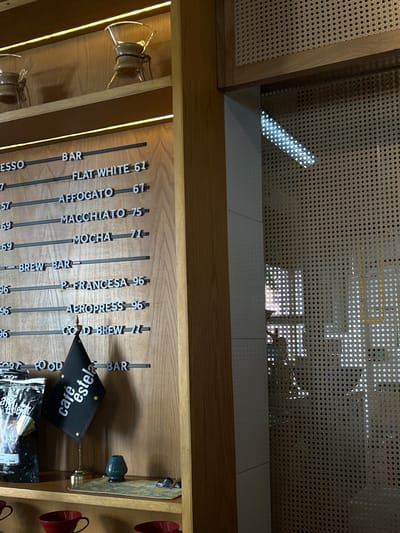 This media file presents an interior view of a cafe in Mazatlán, Mexico, focusing on a display area or counter. No people are visible. The scene is dominated by a vertically arranged menu board crafted from wooden or metal strips, illuminated by integrated strip lighting.
The menu lists various coffee beverages and their prices, likely in Mexican Pesos. Visible items include "FLAT WHITE 61", "AFFOGATO 67", "MACCHIATO 75", "MOCHA 71", "PRENSA FRANCESA 96" (identified as "P FRANCESA" by OCR), "AEROPRESS 96", and "COLD BREW 77". Categories like "BAR", "BREW BAR", and "FOOD BAR" segment the offerings. A small, black flag-shaped sign with white text reads "cafestela", identifying the establishment.
To the left, several clear glass coffee-making devices, possibly pour-over drippers or carafes, are displayed on lit shelves. On the right, decorative red coffee mugs hang on hooks, and a small green ceramic knob is visible.
Above the display, a large woven cane panel forms part of the interior design, suggesting a natural and rustic aesthetic. Bright, natural light streams through a window or opening behind this panel, casting distinct geometric patterns of light and shadow through the cane weave onto the light-colored floor, indicating it is daytime. The overall setting is clean, modern, and inviting, with a focus on warm wood tones and interesting textures.