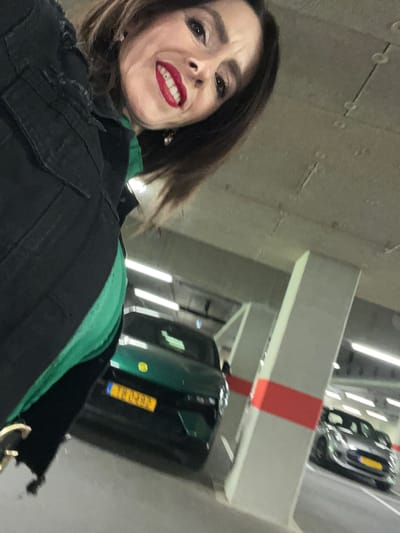 The image, rotated 90 degrees clockwise, captures a smiling woman in what appears to be an underground parking garage. The woman, with brown hair, is wearing a black jacket over a green top and has bright red lipstick. A dental appliance, possibly braces or a retainer, is visible on her lower teeth as she smiles widely, looking directly at the camera.
The background shows elements of a modern concrete parking structure. To the left of the woman, a dark green car is visible, displaying a yellow European license plate with a blue strip on the left. The characters "YX-425" are clearly discernible on the license plate, with two more blurry characters following, consistent with a Luxembourgish registration.
The garage features concrete beams and pillars, with a prominent red stripe visible on one of the beams. Bright, rectangular fluorescent lights illuminate the space. Another vehicle, possibly a silver or grey car, is partially visible in the distance, suggesting multiple parking levels or a car lift system. The overall setting indicates an indoor parking facility, likely in Mensdorf, Luxembourg, given the context and license plate details. The lighting suggests it's an artificial environment, so time of day is not indicated.