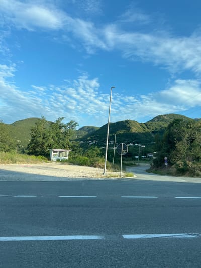 The image depicts a roadside scene in Markovići, Montenegro, characterized by a paved road with white lane markings in the foreground. To the left of the road, there is a gravel area with a small, white, shed-like structure featuring windows and a pink trim. A tall, slender lamppost stands near the gravel area. Beyond the road, a winding asphalt road leads into a landscape of lush green, tree-covered hills. Scattered across the hillsides are small clusters of houses. Several road signs are visible: a hexagonal black sign with white text is positioned near the lamppost, and circular signs indicating speed limits of 70 and 40 are placed further along the winding road. The sky is a vibrant blue, dotted with scattered white clouds. The environment appears to be rural or semi-rural, with a focus on natural topography and infrastructure.