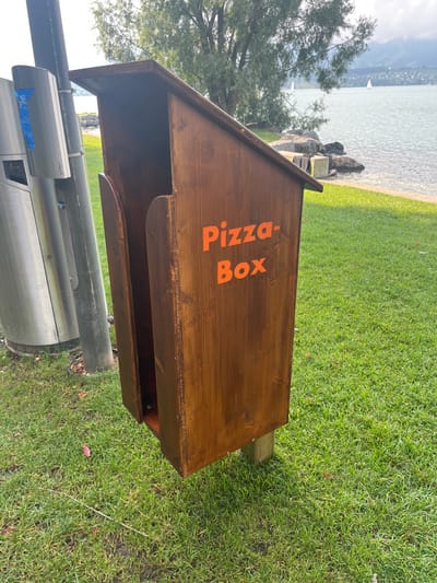 The image presents a dedicated wooden "Pizza-Box" disposal unit situated on a grassy lakeside embankment in Hünibach, Switzerland. The dark brown, wood-stained rectangular box features a sloped roof and an orange, vertically aligned "Pizza-Box" inscription on its front panel. A narrow, vertical opening on the left side of the unit is designed for inserting flattened pizza boxes. The unit is elevated on a visible wooden post embedded in the green grass. To its immediate left, a tall silver pole and a metallic cylindrical waste bin with blue markings are partially visible. The background features a large body of water, likely Lake Thun, with a rocky shoreline separating it from the grass. Across the lake, distant hills or mountains are discernible, and two white sailboats are present on the water. A tree with dark green foliage stands behind the disposal unit. The scene is illuminated by bright daylight, casting clear shadows. No individuals are depicted.