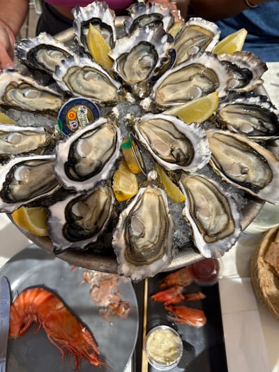 The image shows a close-up, eye-level view of a platter of fresh oysters served on crushed ice, garnished with lemon wedges. A small, round tin labeled "BEURRE D'ISIGNY" is nestled among the oysters. The platter appears to be made of metal, and it is placed on a table. In the foreground, to the left, a grey plate holds a large, cooked prawn and a knife. To the right, a black tray contains several cooked prawns, two small plastic containers of sauce (one red, one creamy), and a basket of bread. The scene suggests a meal at a restaurant. The presence of oysters and the coastal setting are indicative of Lège-Cap-Ferret, France, a region known for its oyster farming and seafood. A person's hand with a red bracelet is visible in the upper left corner, holding the edge of the platter.