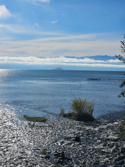 This is a landscape photograph of a body of water with mountainous terrain in the background. To the right of the image, there's a rocky shoreline with some vegetation. A long pier extends into the water. The sky is partially covered with clouds. The water surface reflects sunlight, creating a sparkling effect. Given the description, the location is likely a lakefront in Villarrica, Chile.