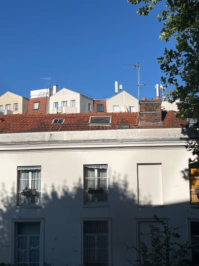 The image depicts the facades of several buildings, observed from a slightly elevated perspective. The sky is clear and bright blue. On the rooftops, several structures are visible, including red tile roofs, dormer windows, chimneys, and various antennas and communication equipment.
In the foreground, the upper portion of a building with a white facade is prominent. This facade features several windows, some with planters containing red flowers. Shadows from trees are cast across the white wall. Below the prominent white facade, another section of a building is visible, with more windows, some of which are barred.
The architectural style suggests an urban setting, likely in a European city. Given the context of L'Haÿ-les-Roses, France, this scene is likely from that locality. No people are visible in the image. The overall impression is one of residential buildings under a clear, sunny sky.