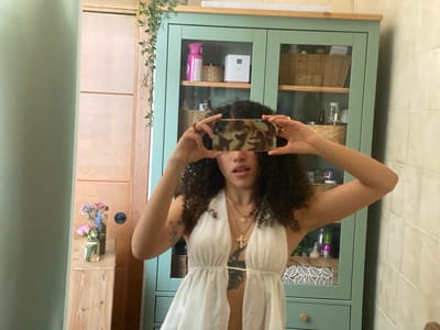 This media file depicts a young person with curly dark hair taking a selfie in a bathroom. The person is wearing a white halter-neck dress and several necklaces. They are holding a smartphone in landscape orientation to take the photo, obscuring their face. Behind them is a light green cabinet with glass doors, revealing shelves stocked with various items, including baskets, bottles, and boxes. To the left of the cabinet, a wooden drawer unit holds a small vase with flowers. The walls appear to be tiled, with a wooden door visible to the far left and right. The overall impression is that of a personal, casual moment captured in a home bathroom, possibly during the daytime given the lighting.