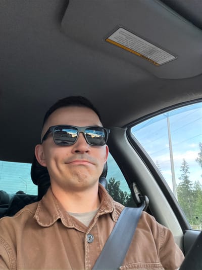 Here is a description of the image:
A man wearing sunglasses and a brown collared shirt sits in the driver's seat of a vehicle. He has his seatbelt on. The car has a sunroof. There is a warning sticker on the sun visor. The surrounding environment is visible through the car windows and sunroof, showing trees and the sky. The picture may have been taken while the car was stationary. There is no indication of Severnyy, Russia in the image.