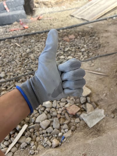 In Rosice, Czechia, a person wearing a light grey work glove with a blue trim on the wrist gives a thumbs-up. The glove is slightly damp. The background shows a construction or renovation site, with loose gravel, broken concrete pieces, rebar, and some orange safety markers on the ground. The lighting suggests it is daytime, likely outdoors. The focus is on the gloved hand and its gesture.