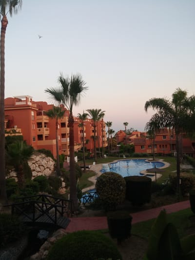 The image depicts a twilight scene at a resort in Artola, Spain. The foreground features a dark wooden footbridge spanning a shallow, dark body of water. Beyond the bridge, a swimming pool with blue water is visible, surrounded by manicured grass and several tropical plants, including palm trees. In the mid-ground, there are a few white lounge chairs and thatched umbrellas. The background is dominated by a row of orange apartment buildings with balconies and white railings. The sky is a pale, gradient hue, transitioning from light blue to a soft pinkish-orange, indicative of sunset or sunrise. A single bird is seen in flight in the upper left portion of the sky. The overall impression is that of a tranquil, tropical vacation setting.
