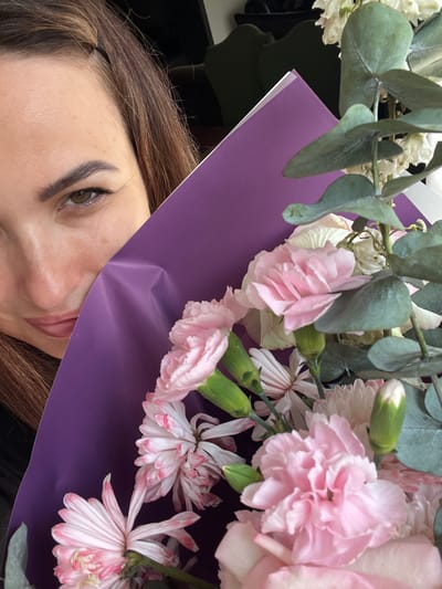 In this close-up, eye-level shot, a young woman, partially obscured by a bouquet of pink and white flowers, is looking towards the camera. Her brown hair is pulled back with a dark band, and she has light brown eyes, dark eyebrows, and fair skin with some light freckles. She is wearing dark clothing. The bouquet, wrapped in vibrant purple paper, features carnations, eucalyptus leaves, and other delicate white and pink blossoms. The background is dark and out of focus, suggesting an indoor setting. The lighting appears to be natural, highlighting the textures of the flowers and the woman's skin.