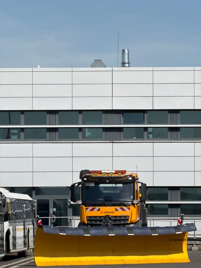 In Balice, Poland, a bright yellow snowplow truck with a large plow is parked in front of a modern white building. The building has multiple horizontal rows of windows. To the left of the snowplow, a white bus is visible. The sky is clear and blue.