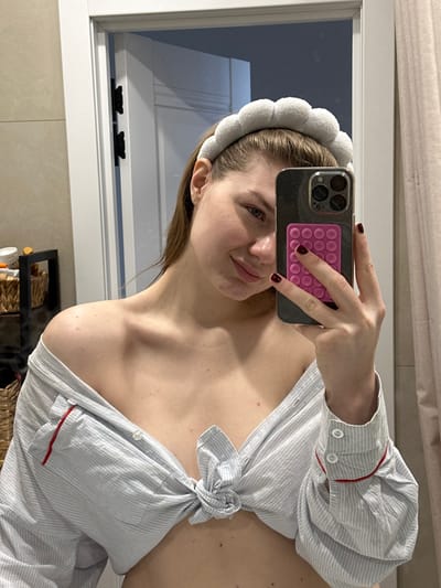A young adult female is depicted in a mirror selfie, located in what is described as the city of Grebeni, Russia, within an indoor setting resembling a bathroom or dressing area. She has fair skin, light brown hair pulled back by a grey, cloud-textured headband, and a slight smile. She wears a light blue and white striped, long-sleeved button-up shirt, unbuttoned and tied at the midriff, exposing her abdomen and shoulders. The shirt cuffs feature red trim. Her left hand holds a dark-colored smartphone, equipped with a bright pink, rectangular textured grip on its back, positioned to capture her reflection. Her fingernails are painted dark red. In the background reflection, a light-colored door with dark hinges is visible to her right. To her left, a dark-framed shelving unit holds various items, including a wicker basket and several containers. The walls are a light, neutral color.