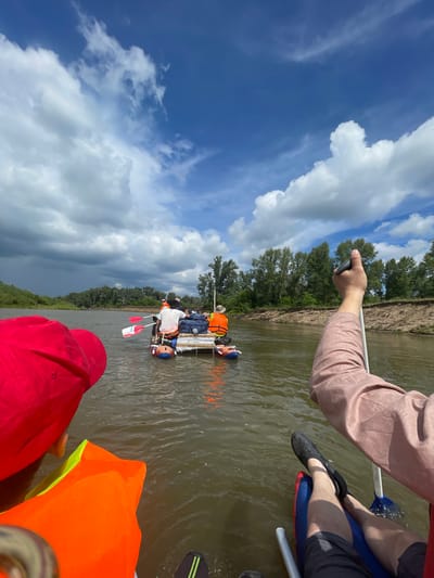 A group of people are on a raft on a river, with a shoreline of trees and a sandy bank in the background. The sky is partly cloudy with blue patches. The raft has several people on it, wearing life vests and holding paddles. One person in the foreground is wearing a red hat and an orange life vest. Another person in the foreground has their arm raised, holding a paddle. The scene appears to be in Samorodovo, Russia.