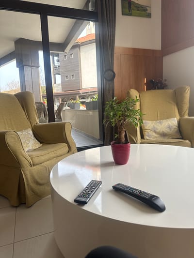 The image shows an interior setting in Bozhurets, Bulgaria. Two light-green upholstered armchairs are visible on either side of a round white coffee table. On the table, there is a potted plant with green foliage and two black remote controls. A large window with a dark frame reveals an exterior view of buildings, possibly residential, across a small courtyard. The image is taken at an angle, giving a tilted perspective of the scene. A golf club set is situated near a window.