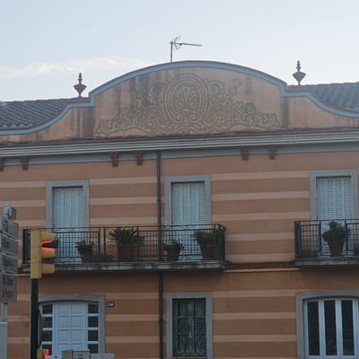 The image shows the facade of a building in El Perelló, Spain. The building is painted in horizontal stripes of a peachy orange color. The upper part of the building features a decorative frieze with a central ornamental motif. There are two balconies with black wrought-iron railings and potted plants. Below the balconies, there are several windows, some with light blue shutters and others with ornate metal grilles. A traffic light with a red light illuminated is visible on the left side of the image, along with street signs. A small black object, possibly a drone or a bird, is in the sky.