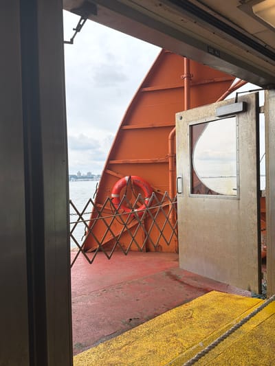 A ferry door, with a glass window, opens onto the deck of a ferry. The deck is painted red and yellow. A life preserver hangs on an orange metal wall. In the distance, a cityscape is visible across the water. The ferry appears to be in the city of Bayonne, United States of America, indicated by a sign that reads "SIE. NJ."