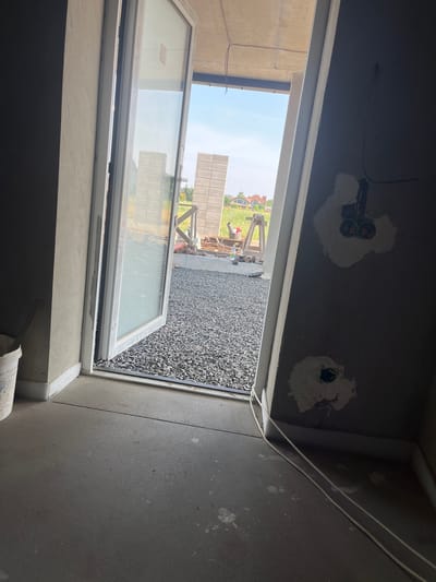 The image shows an open doorway leading to a construction site. The interior of the building is partially finished, with concrete floors and gray walls. Electrical outlets are visible on one of the walls, with wires running along the floor. Outside, there is a gravel-covered area with stacks of building materials and a partially constructed wall. In the distance, houses and trees can be seen under a clear sky. The scene suggests an ongoing construction project in a suburban or rural setting.
