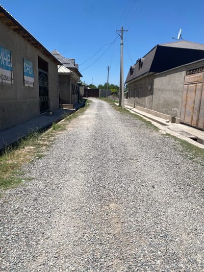 The image shows a gravel road leading into a village in Bogiston, Tajikistan. On the left, a building with a gray facade has blue signs with white text, and a metal gate. Further down the road, there are more buildings with similar gray facades, some with tiled roofs, and a large, rusted metal gate. On the right, another building with a gray facade and a dark tiled roof features a satellite dish. A utility pole with wires stands on the right side of the road, and trees are visible in the background. The sky is clear and blue.