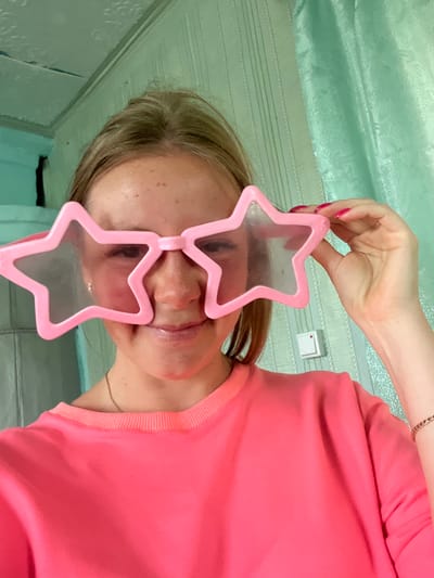 The image shows a close-up of a person wearing large, pink star-shaped novelty glasses. The person's face is visible, with their eyes looking through the star-shaped lenses. They are wearing a bright pink top. The background appears to be a room with light green patterned wallpaper and horizontal lines, possibly suggesting a ceiling or a wall. A white light switch is visible on the wall. The lighting suggests it is daytime. The overall mood is playful and lighthearted due to the novelty glasses. There is no visible text in the image.