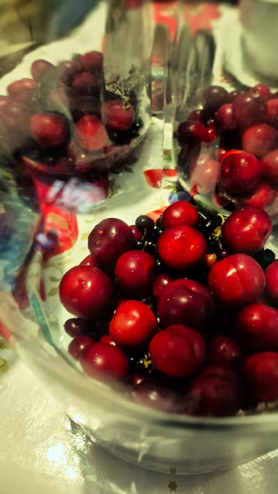 The image shows a close-up, top-down view of a clear plastic bag filled with red cherries and some black currants. The bag is partially transparent, revealing the textured surface of the bag itself and the shiny, round fruits inside. The cherries are a vibrant red, with some appearing darker and more purple-hued. They are clustered together, with a few black currants mixed in. Some of the fruits have small droplets of water on their surfaces, suggesting they may have been recently washed. The lighting is somewhat dim and diffused, creating soft highlights and shadows on the fruits. The background is blurred and indistinct, but appears to be a light-colored surface, possibly a countertop or table. The overall impression is one of fresh produce, likely prepared for consumption or storage. There is no visible text in the image, nor any indication of a specific time of day or weather conditions. The setting is implied to be domestic or possibly a market stall, given the nature of the contents.