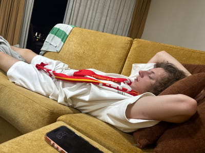 A young person is asleep on a mustard-colored couch, with their head resting on a brown pillow. They are wearing a white t-shirt and white shorts, with a red and yellow scarf draped over their chest. A grey and green striped towel is draped over the back of the couch. A smartphone is visible on the couch in the foreground, displaying the time 8:41. Through a window in the background, lights from a city at night can be seen. The scene appears to be indoors, likely in a residential setting within the city of Al Fiqait, United Arab Emirates.