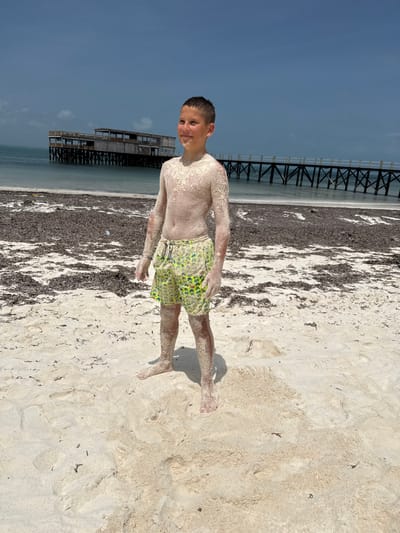 A young, shirtless male, approximately pre-teen, stands smiling on a white sandy beach in Michamvi Kae, Tanzania. His torso, arms, and legs are significantly coated in light-colored sand. He wears yellow and green patterned swim shorts. Behind him, the beach transitions to a broad band of dark brown, dried seaweed or vegetation at the water's edge. Beyond this, light blue-green ocean water is visible. In the distant background on the left, a multi-level wooden structure built on stilts stands over the water. To the right of this building, a long, dark wooden pier extends into the ocean from the shore. The sky is a light blue with darker, cloudier regions towards the horizon.