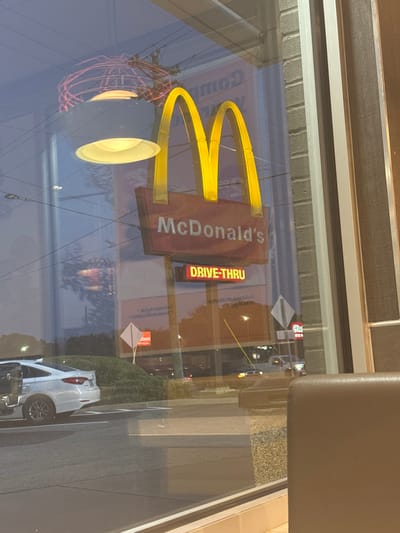 A McDonald's sign with golden arches and "DRIVE-THRU" is reflected in a window. Outside the window, a white car is parked near a green bush. Streetlights and possibly other signs are visible in the reflection. A portion of a building's interior, including a chair, is visible in the upper right corner. The scene is captured through the glass window.