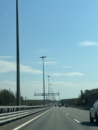 The image shows a highway in Pesochny, Russia. The view is from a vehicle on the rightmost lane, looking straight ahead. The road is lined with metal guardrails and tall light poles with anemometers. Several cars and trucks are visible in the distance. The sky is blue with scattered white clouds. The road is surrounded by trees.