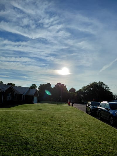 The image depicts a bright, sunny day in Berea, United States of America. The scene is a suburban residential area, characterized by a neatly manicured green lawn in the foreground that slopes gently upwards. Beyond the lawn, a row of houses with dark metal roofs and brick exteriors is visible. A paved driveway runs alongside the lawn, and several dark-colored vehicles are parked along it. Mature trees with dark green foliage line the property, casting shadows on the driveway and houses. The sky is a vibrant blue, crisscrossed with wispy white clouds. The sun is high in the sky, its bright light creating a lens flare and casting a warm glow over the scene. The overall impression is one of a peaceful, pleasant summer day. The image appears to have been taken in the late afternoon or early evening, given the angle of the sun and the warm lighting. There is no discernible activity or event taking place, as the scene appears static and unoccupied. There is a red stop sign visible near the parked cars.