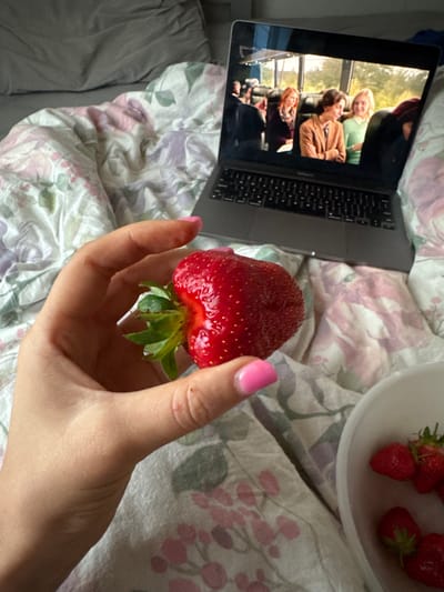 The image presents a cozy, indoor scene from a first-person perspective, focused on relaxation and simple pleasures. In the foreground, a hand with pink manicured fingernails holds a large, vibrant red strawberry, showcasing its texture and green stem. To the upper right, a white bowl filled with several more fresh strawberries is partially visible, indicating a ready supply.
A silver/grey laptop, resembling a MacBook, is positioned on the left side of the frame, open and displaying content on its screen. The screen shows a scene from what appears to be a movie or TV show. Three individuals are prominently featured: a blonde woman in a light green top smiling broadly, a person with brown hair in a brown jacket laughing, and another person with reddish hair partially visible in the background. They appear to be inside a vehicle, possibly a bus or train, suggested by the window frames and glimpses of greenery outside.
The setting is a relaxed personal space, likely a bedroom. The laptop and strawberries are resting on bedding. A floral-patterned duvet or blanket with intricate designs in shades of pink, green, and purple on a white background covers the bed. A solid dark grey sheet or blanket is also visible on the far left. The overall activity depicted is leisurely, combining watching entertainment with enjoying fresh fruit. No specific text is clearly visible on any objects within the image. The scene conveys a sense of comfort and unwinding. The location of Fehraltorf, Switzerland, is not discernible from the visual cues in the image.
