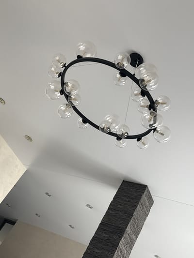 The image shows a modern, elliptical chandelier with multiple clear glass globes suspended from a black metal frame. The chandelier is mounted on a white ceiling. The ceiling also features several recessed circular lights and a few square light fixtures. In the lower part of the image, a textured, dark gray rectangular column extends upwards towards the ceiling. The lighting suggests an indoor environment, possibly a residential or commercial space. The image does not contain information to determine the location in the city of Barysaw, Belarus.