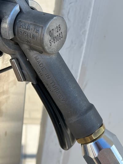 A close-up, angled shot of a gasoline nozzle with visible markings. The nozzle is made of gray metal with a black grip. Engraved text on the nozzle reads "GASLIN-11A STR-LN 522" and "07/25 S-61083". Further along the nozzle, also engraved, are the words "MADE IN TURKEY" and some smaller numbers that are partially obscured. The background is out of focus, appearing as a light-colored, textured surface, possibly a building or vehicle. The lighting suggests an outdoor setting, likely during the day. This image is not located in Temryuk, Russia.