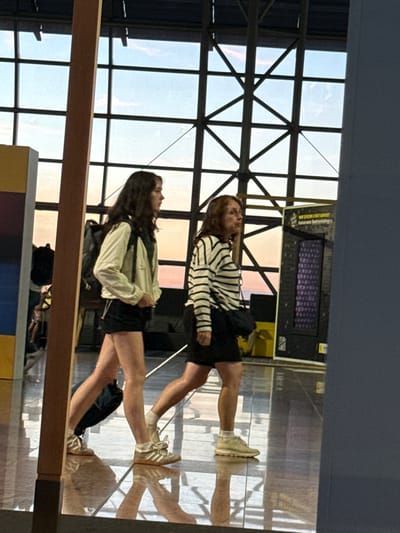 Two individuals, a younger woman with dark hair and an older woman with auburn hair, are walking through Brussels Airport in Zaventem, Belgium. The younger woman is on the left, wearing a light-colored jacket, a dark t-shirt, and black shorts, carrying a black backpack. The older woman is on the right, wearing a striped long-sleeved shirt and a black skirt, with a dark crossbody bag. They are walking on a polished floor that reflects the ceiling and the window structure behind them. The background features a large window with a grid-like metal structure, revealing a sky with soft, diffused light, possibly indicating dusk or dawn. To the right of the older woman, a digital display board is visible. The overall scene appears to be a transit area within the airport.