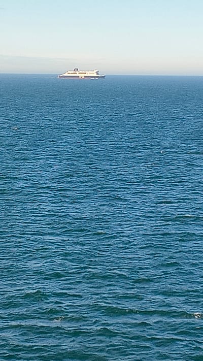 A ferry boat is visible in the distance on a calm, blue sea under a clear sky. The boat is white with orange and black lettering. The water has small ripples and is a deep blue. The horizon is visible in the distance, a faint line separating the sea and sky. There are no people or other significant objects visible in the immediate foreground or on the boat.
