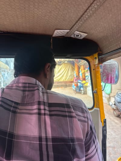 This image depicts a view from inside an auto-rickshaw, looking forward onto a street scene in Gokarna, India. In the immediate foreground, the back of a person's head and right shoulder are visible, with the individual wearing a light-colored plaid shirt featuring white, grey, and black checks. Above, the beige, textured interior ceiling of the auto-rickshaw is present, crossed by a patterned strap. An Ace of Clubs playing card is secured to the ceiling by this strap. The auto-rickshaw's yellow metal frame and a side-view mirror are visible on the right.

Through the vehicle's windows, a street or lane is observed. On the left, a structure is partially covered by a large yellow tarpaulin. Towards the center-right, colorful textiles or goods are displayed on what appears to be market stalls. A person wearing a reddish-pink top is standing near a parked light-blue scooter in this area. Another grey vehicle, possibly a scooter or motorcycle, is partially visible on the far right, both through the window and reflected in the side mirror. The environment suggests a commercial or market street within Gokarna.