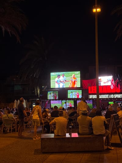 The image captures a lively outdoor scene at night, likely in Playa De Las Américas, Spain, as suggested by the palm trees and warm lighting. A group of people are gathered, seated at tables and standing, all facing multiple large screens displaying what appears to be a sporting event. The screens show close-ups of athletes in uniforms, suggesting a soccer or football match. The atmosphere seems casual and engaged, with individuals engrossed in the game.
The setting is an outdoor dining or bar area, characterized by rows of chairs and tables, and illuminated by warm, ambient lighting from overhead lamps and the glow of the screens. One large screen prominently displays bright red lettering with white accents, and to its right, smaller text reads "OF ENTI TAINMENT AND SPORTS". Above this, there are flags, including what looks like the American flag and possibly another national flag. A maroon flag with a star is also visible on one of the screens. The time of day is clearly night, indicated by the dark sky and artificial lighting. The weather appears to be clear, allowing for an outdoor gathering. The people are dressed casually, suitable for a warm evening. Some individuals are seen holding drinks, further emphasizing the social and relaxed nature of the event. The overall mood is one of shared enjoyment and attention focused on the televised sporting activity.