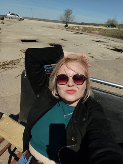 A woman with fair hair and sunglasses is posing for a selfie. She is wearing a black jacket over a teal top and a silver necklace. Her left arm is raised with her hand behind her head. She appears to be sitting on a bench with a black leather backrest and a wooden seat. The location is likely outdoors, based on the background. In the background, there is concrete ground, a small body of water, trees, and two cars. Based on the prompt, the location is Икское Устье, Russia.