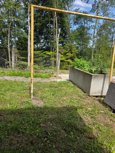 A low-angle outdoor shot shows a weathered concrete planter box filled with lush green plants. The planter is positioned on a grassy slope next to a black asphalt-like surface. A yellow metal frame, possibly part of a sports goal, extends into the foreground, its paint peeling in places. Behind the planter, a chain-link fence delineates a boundary, with trees and foliage visible beyond it. The scene suggests a park or recreational area. The lighting indicates it is daytime, with sunlight casting shadows on the grass. No people or text are visible in the image. The weather appears to be clear and sunny. The overall impression is one of an outdoor, possibly neglected, space with a touch of greenery.